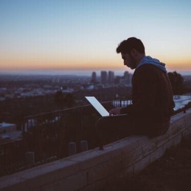 Mann, im Abendlicht, sitzt hoch über der Stadt und arbeitet am Computer.