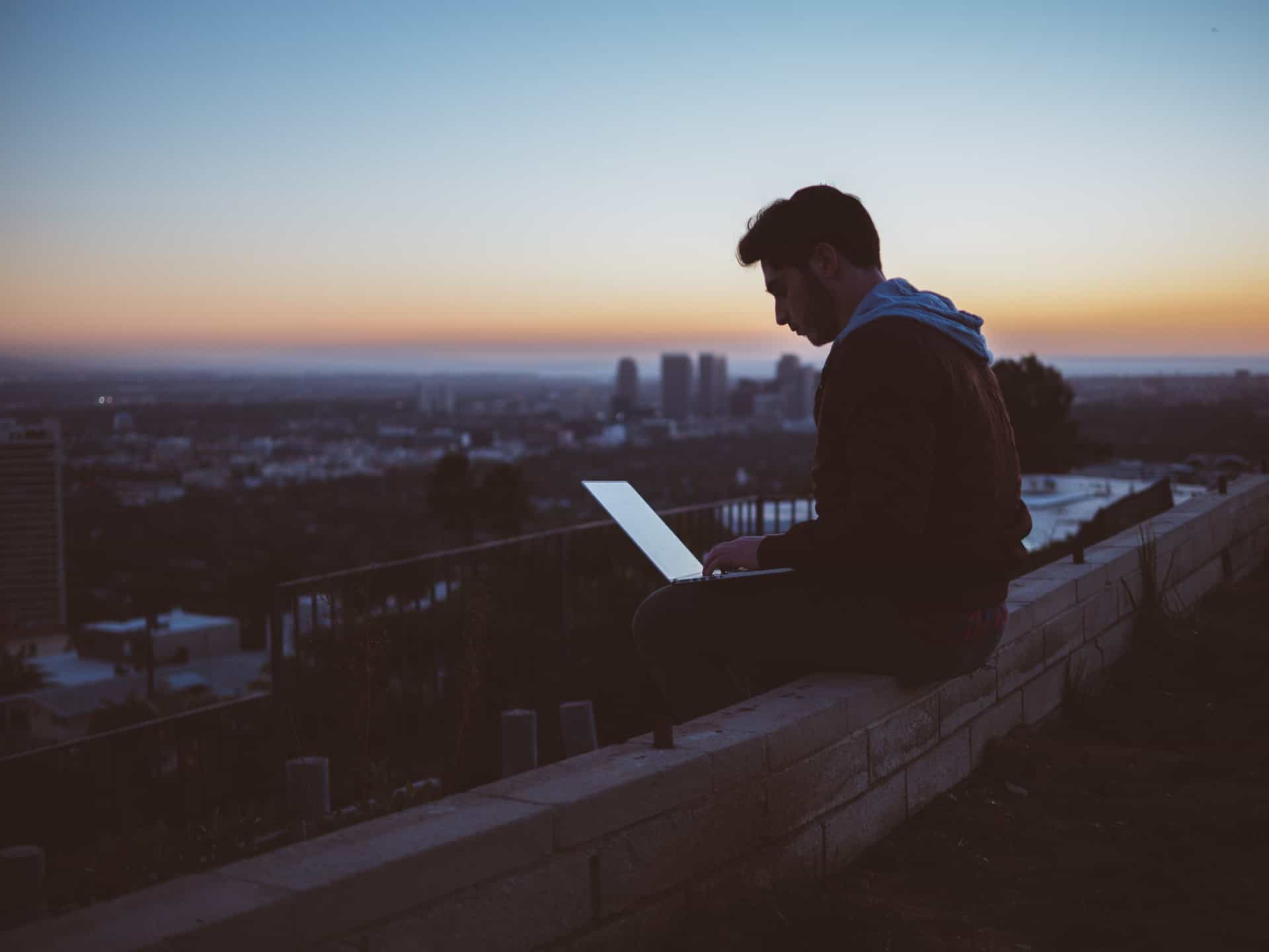 Mann, im Abendlicht, sitzt hoch über der Stadt und arbeitet am Computer.